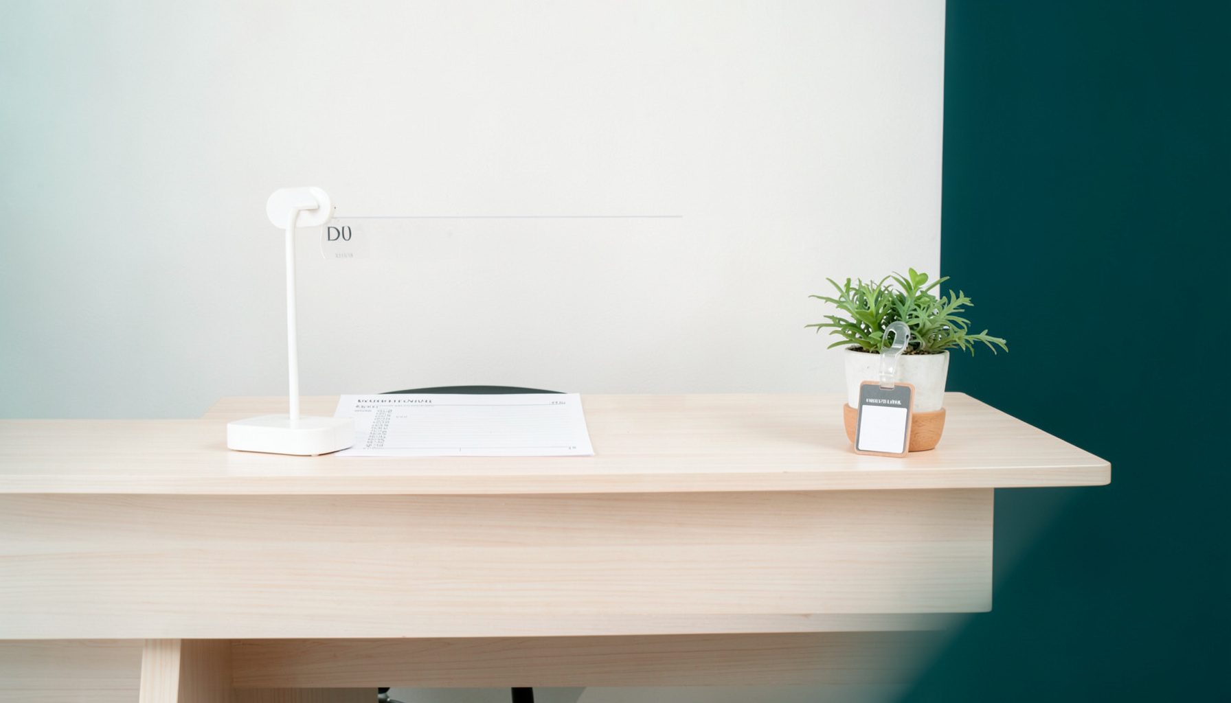 A clean, bright conference registration desk with a neatly arranged sign-in sheet, a name-badge holder, and a small potted plant on a light wood table, set against a soft off-white wall.